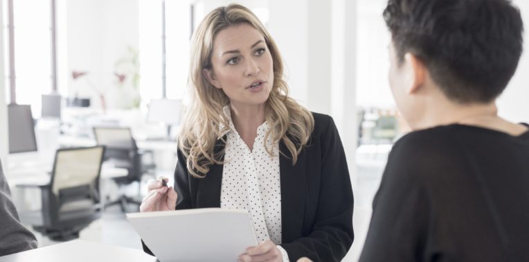 Woman holding papers and a pen talking to another person