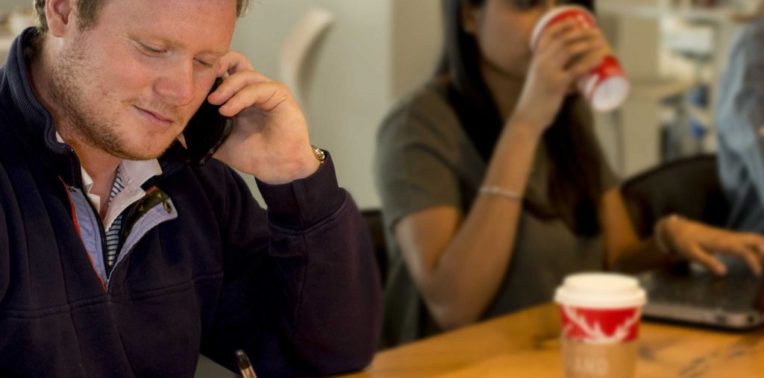Man talking on a cell phone at a table with a woman drinking from a red coffee cup in background