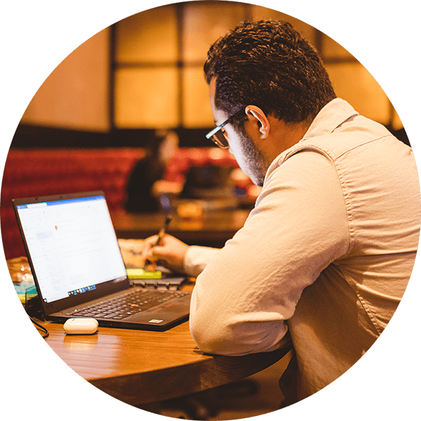 A man sitting at a table with an open laptop while writing on a pad of paper