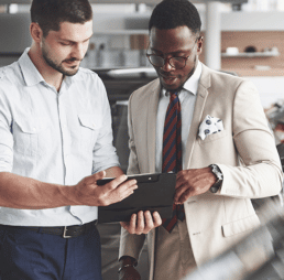 A man in glasses and a suit and a man in a button-up shirt study software on a tablet screen